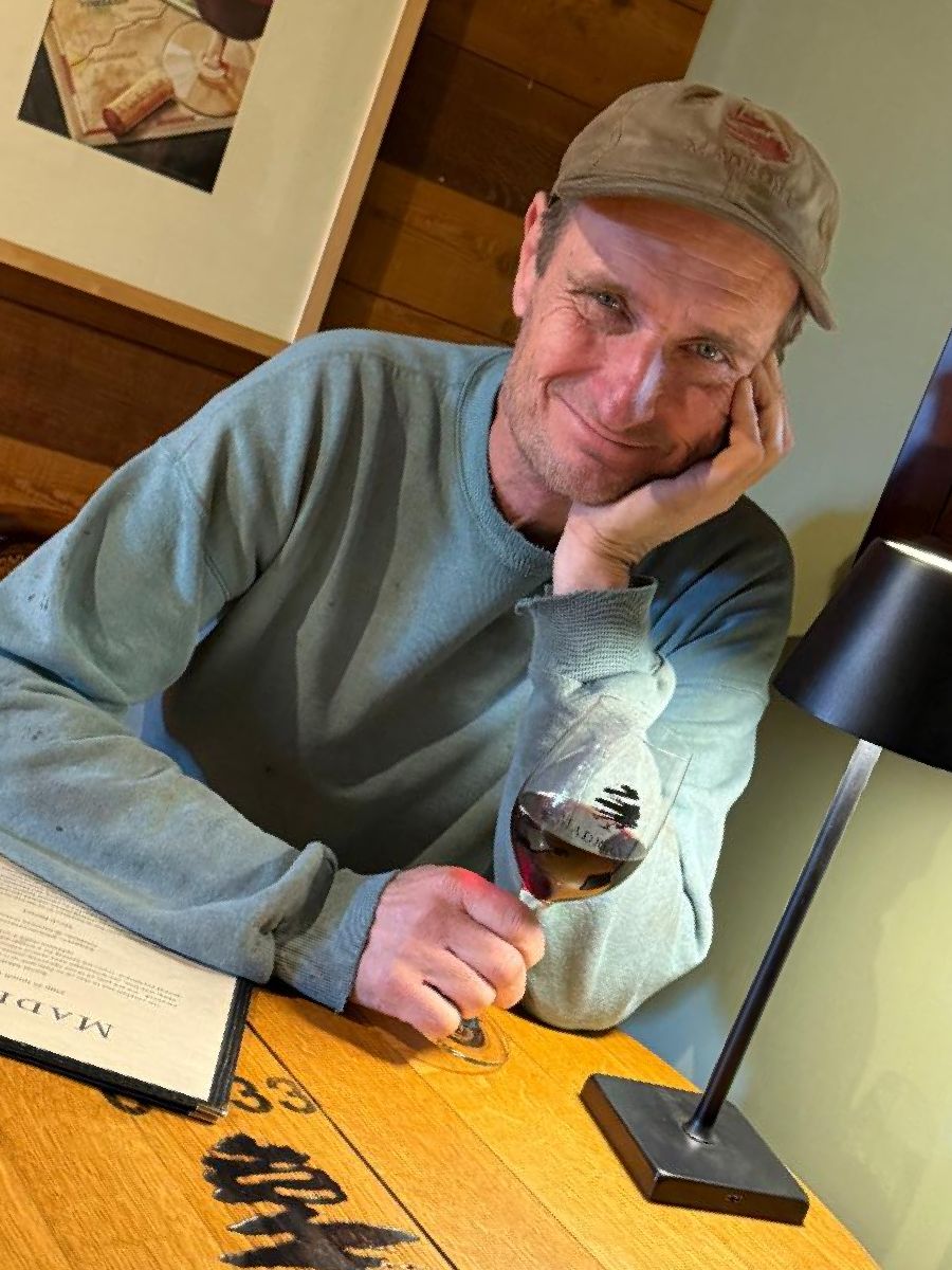 Paul in a cap resting his chin on his hand at a table with a glass of red wine