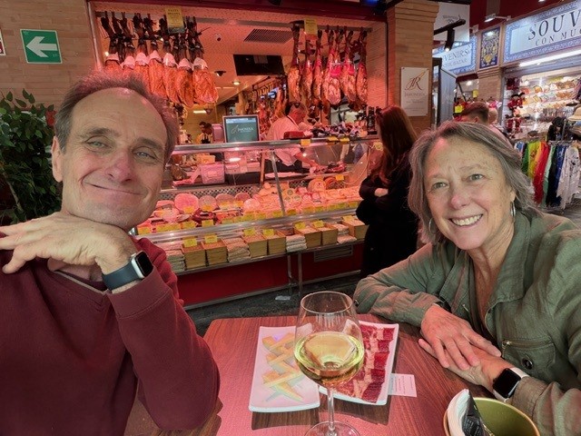 Paul and Maggie at a tapas bar counter with a glass of white wine, cured hams hanging behind them