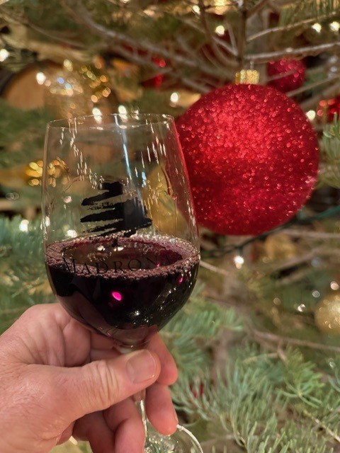 A hand holding a glass of red wine in front of a Christmas tree with red ornaments